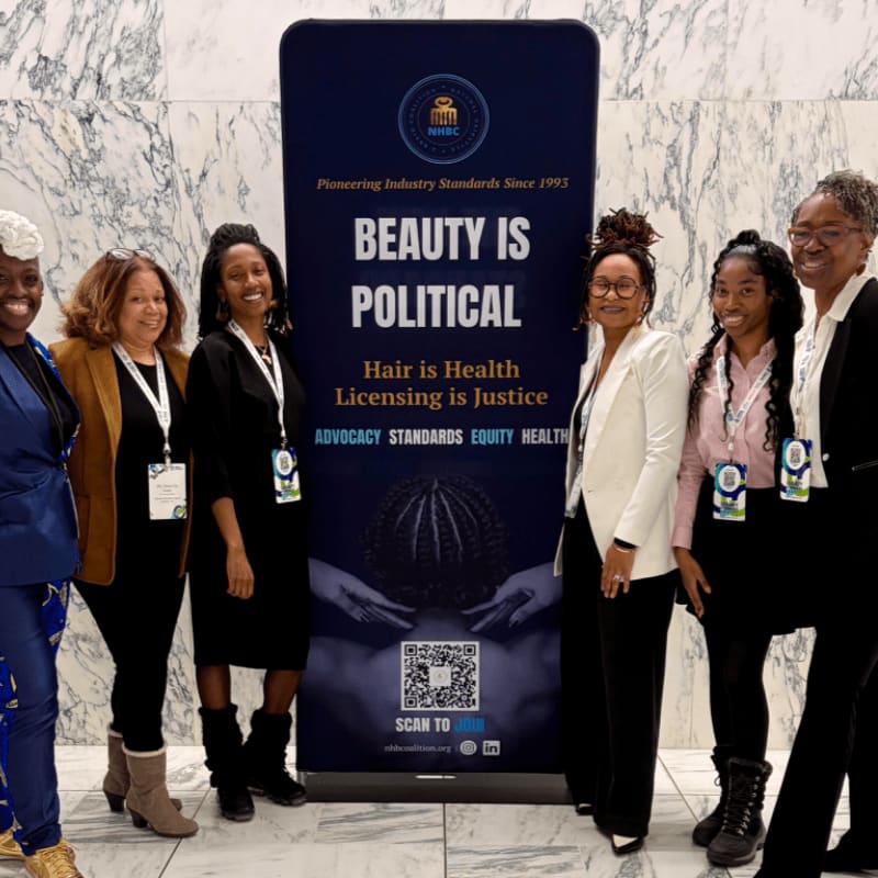 Six women stand in front of a banner that reads "BEAUTY IS POLITICAL" with a marble floor and wall in the background.