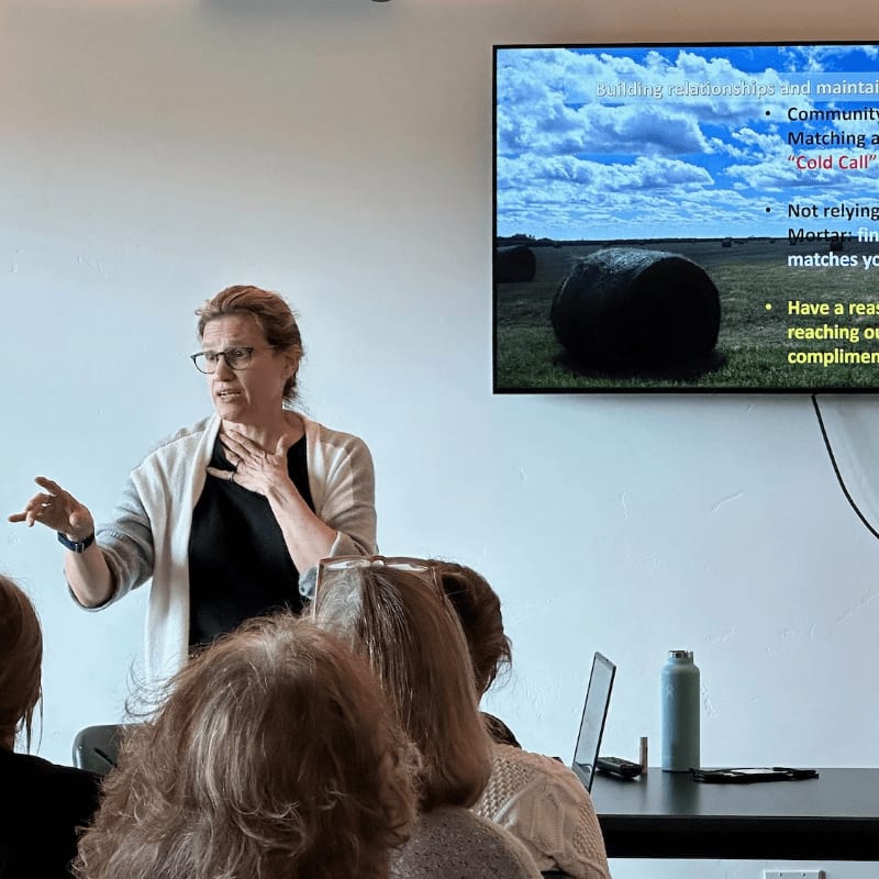A woman gestures while presenting to an audience, with a slide displaying text and an image of hay bales in a field behind her.