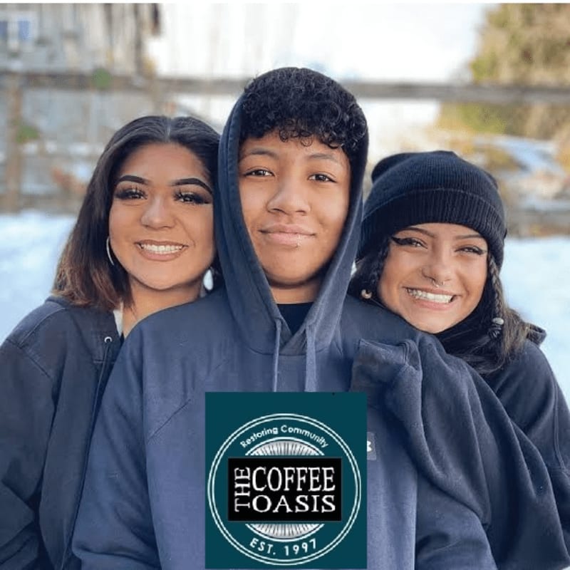 Three smiling young people are posing in front of a snowy background, with a "The Coffee Oasis" logo superimposed on the boy in the center.