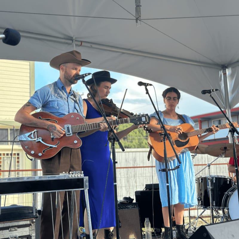 Three musicians play instruments on a stage under a white tent, with buildings and trees visible in the background.