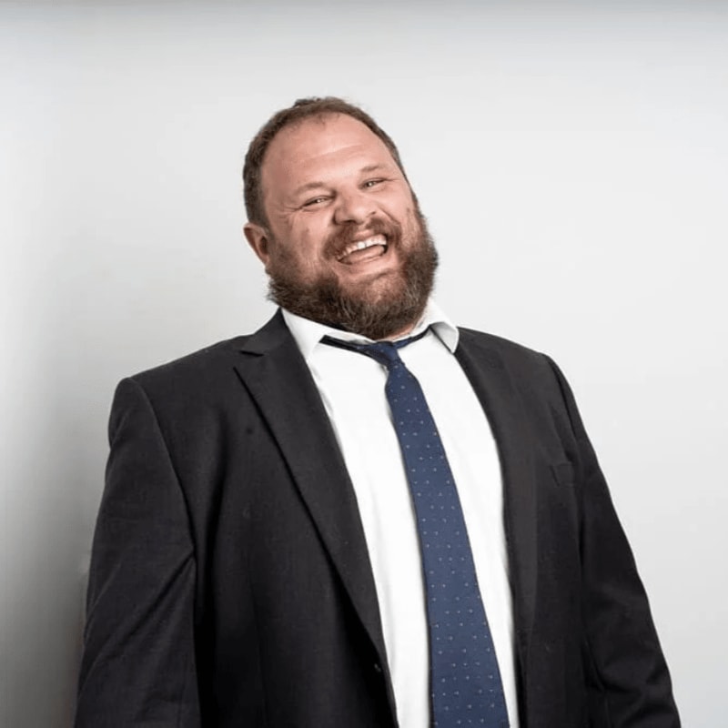 A bearded man in a suit and tie laughs heartily against a plain white background.
