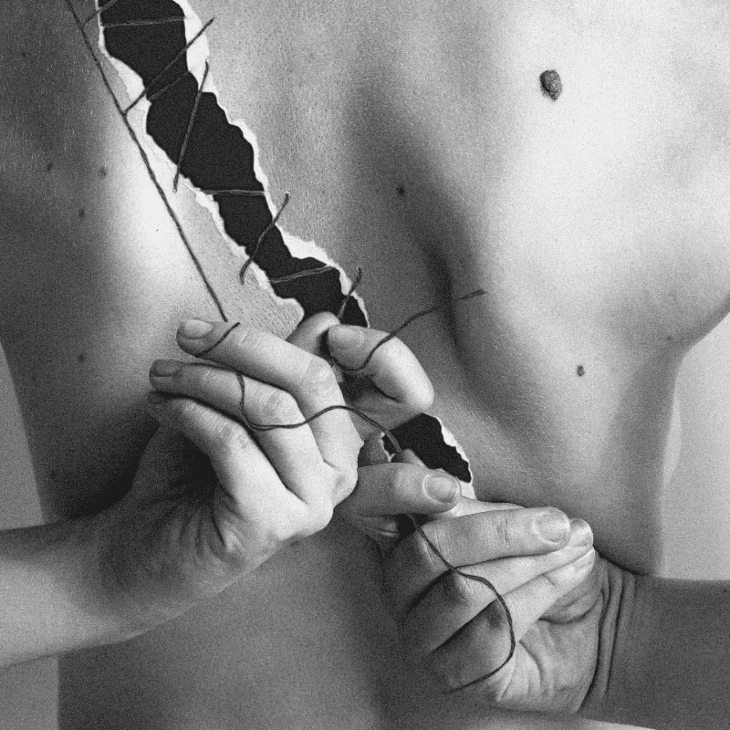 Hands stitch together a torn torso against a plain background.