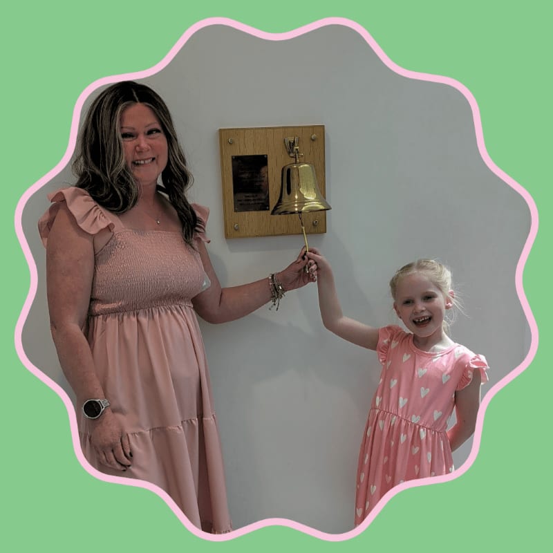 A woman and a young girl in pink dresses smile as they ring a brass bell mounted on a wooden plaque against a white wall, framed by a scalloped pink border on a light green background.