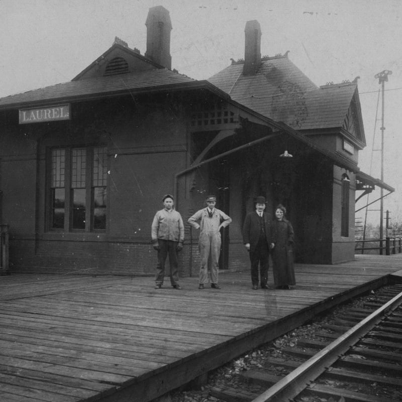 Four people stand on a wooden train platform in front of the Laurel train station, with railroad tracks in the foreground and a windmill visible in the background.