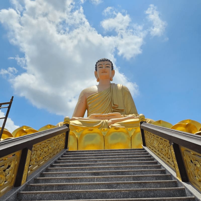 A grand golden Buddha statue sits serenely against a bright blue sky with fluffy white clouds, approached by a wide stone staircase with ornate golden railings.