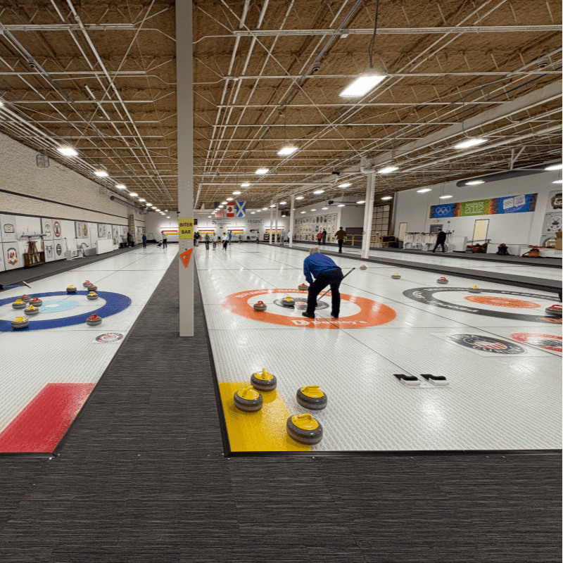 In the foreground, yellow curling stones rest on a yellow mat, while in the background, a person in a blue shirt sweeps the ice on a curling rink with other stones scattered across it.