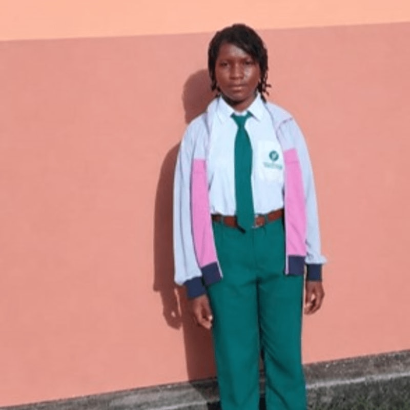 A young girl in a school uniform stands against a peach-colored wall.
