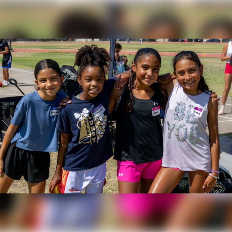 Four smiling young girls pose for a photo in the foreground, with a blurred outdoor track and field in the background.