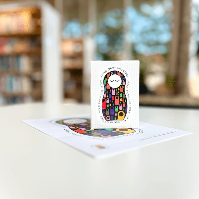 A small card with a nesting doll design sits on a larger print of the same design, with a blurred background of bookshelves and windows.