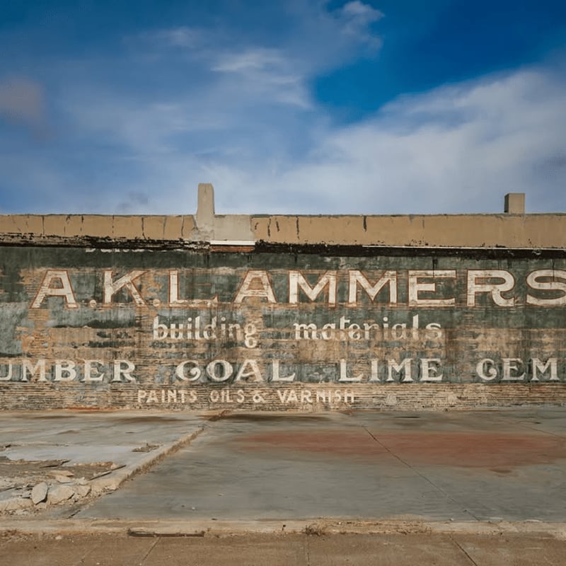 A weathered, faded mural advertising building materials adorns a brick wall in the background, while a cracked concrete sidewalk occupies the foreground under a bright blue sky.