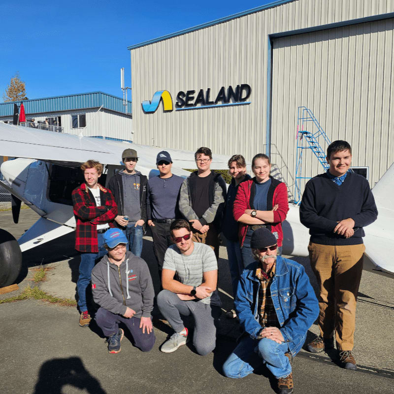 A group of young people poses in front of a small airplane and a building with "SEALAND" signage.