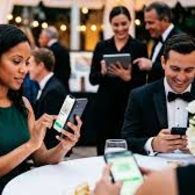 In the foreground, people in formal attire are using their smartphones at a table, while in the background, servers attend to guests at an event.