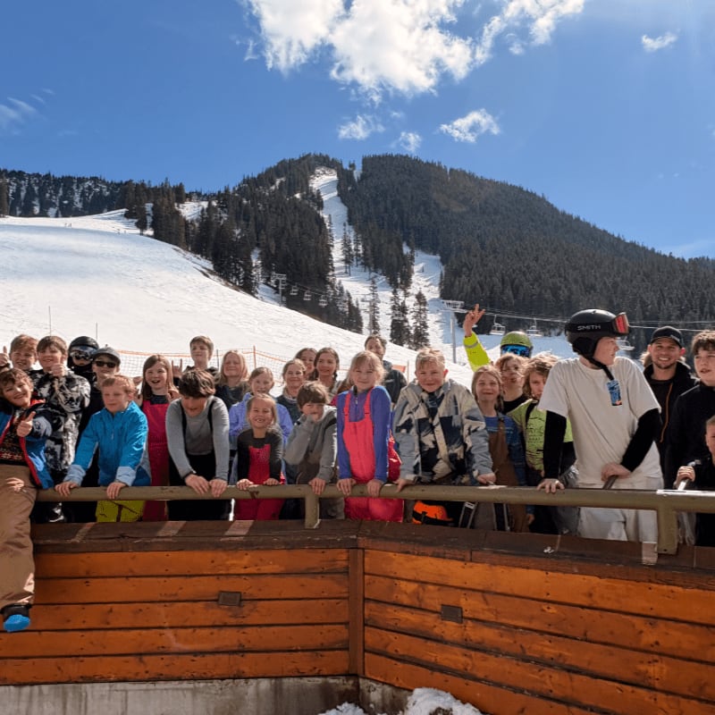 A group of children pose for a photo in front of a wooden railing with a snow-covered mountain and ski slopes in the background.