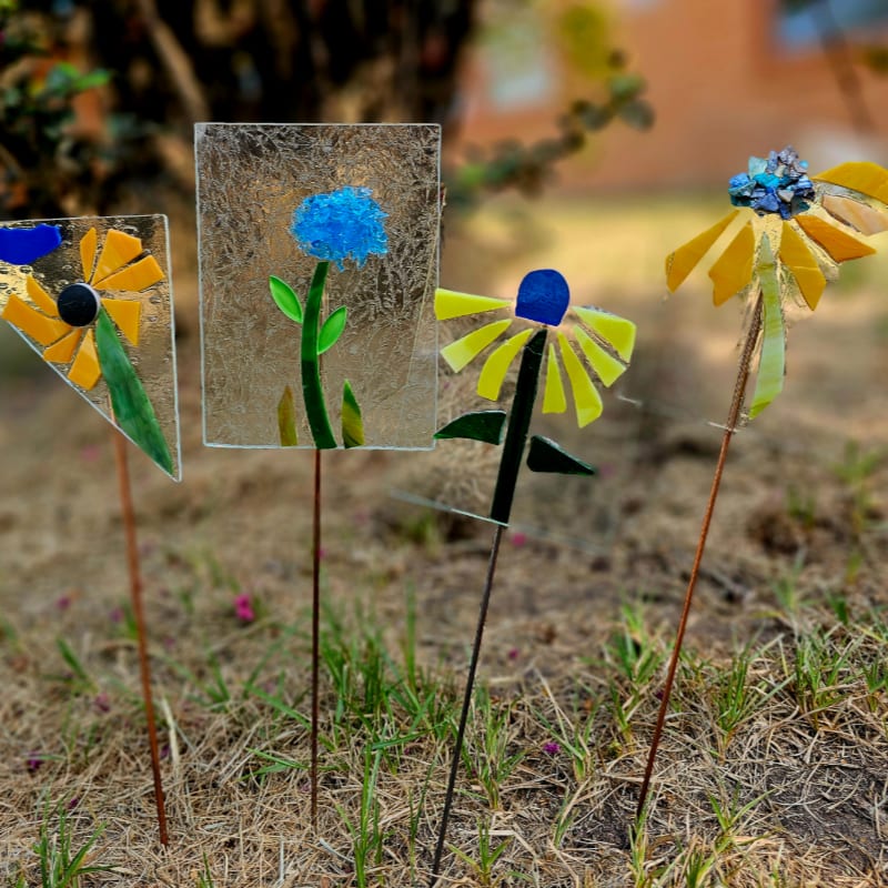 Four colorful mosaic garden stakes, resembling flowers, stand in dry grass with a blurred building in the background.