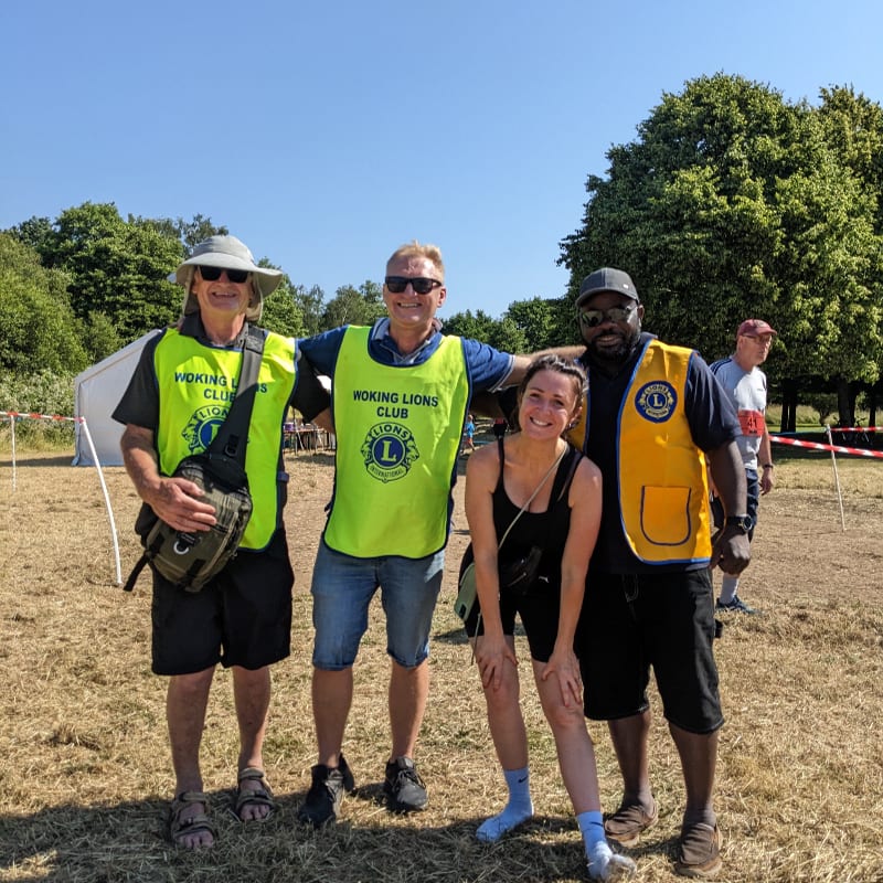 Four people in bright yellow vests stand in a grassy field with trees and a tent in the background.