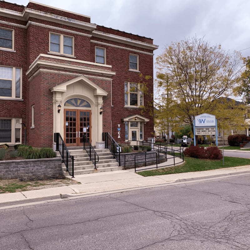 A brick building with a prominent entrance and stairs is in the foreground, while a street and a sign with trees are in the background.