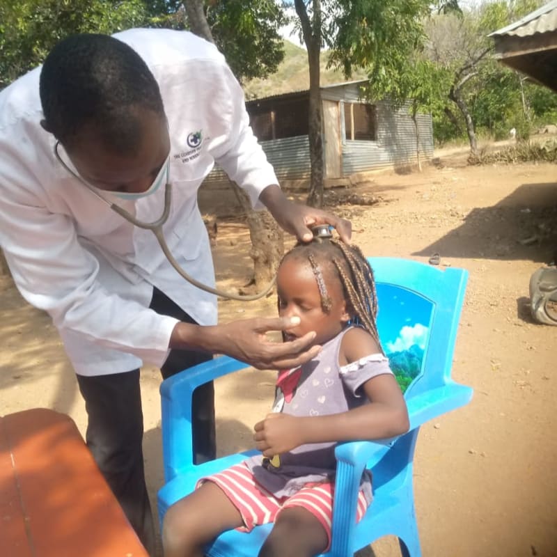 A doctor in a white coat examines a young child sitting on a blue chair in a rural outdoor setting with simple buildings in the background.
