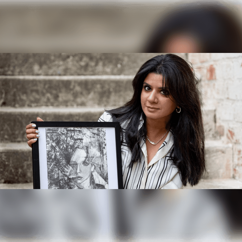 A woman in a striped pajama set sits on concrete steps with a framed picture to her right, against a brick wall.