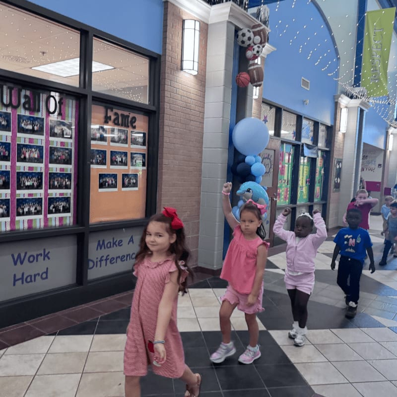 In the foreground, three young girls walk down a hallway, while the background features a "Wall of Fame" and blue walls adorned with sports memorabilia.