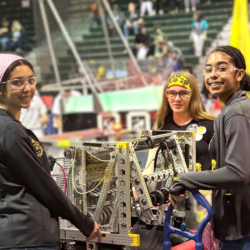 Three young women in safety glasses stand in front of a complex robot, with a blurred audience in the background.