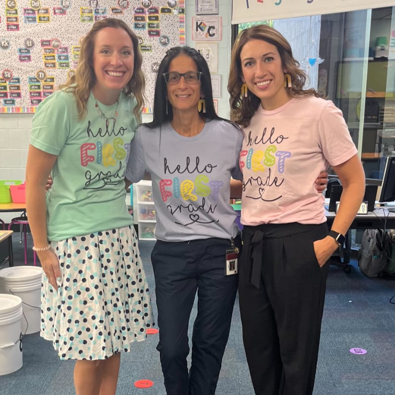 Three smiling women in "hello first grade" t-shirts pose in a colorful classroom with a whiteboard and desks in the background.