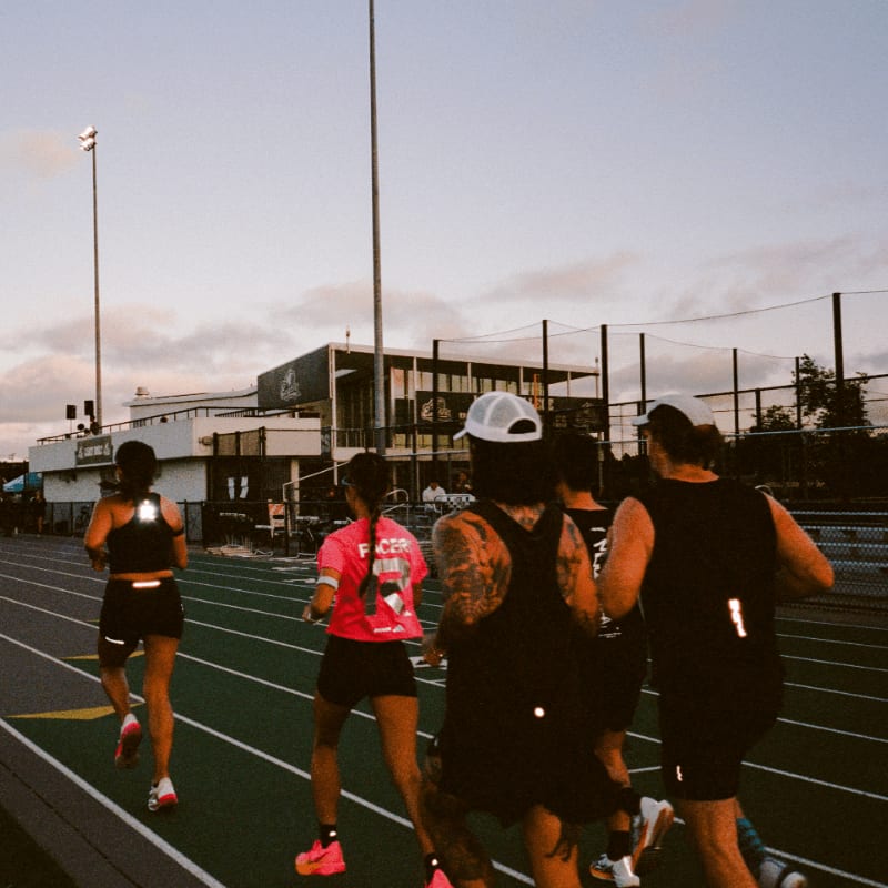 A group of runners are on a track in the foreground, with a building and stadium lights in the background under a cloudy sky.