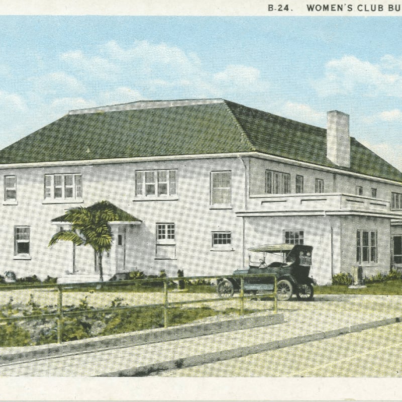 A vintage car is parked in front of a large, light-colored building with a green roof, set against a backdrop of a cloudy blue sky.