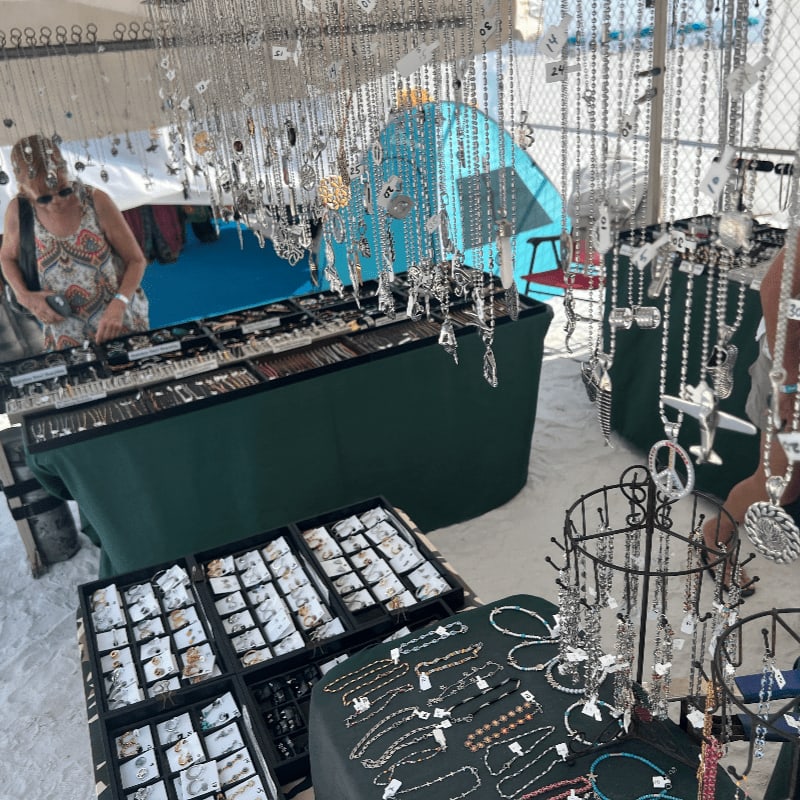 A woman browses a jewelry stall filled with necklaces and bracelets, with a blue tent and a fence visible in the background.