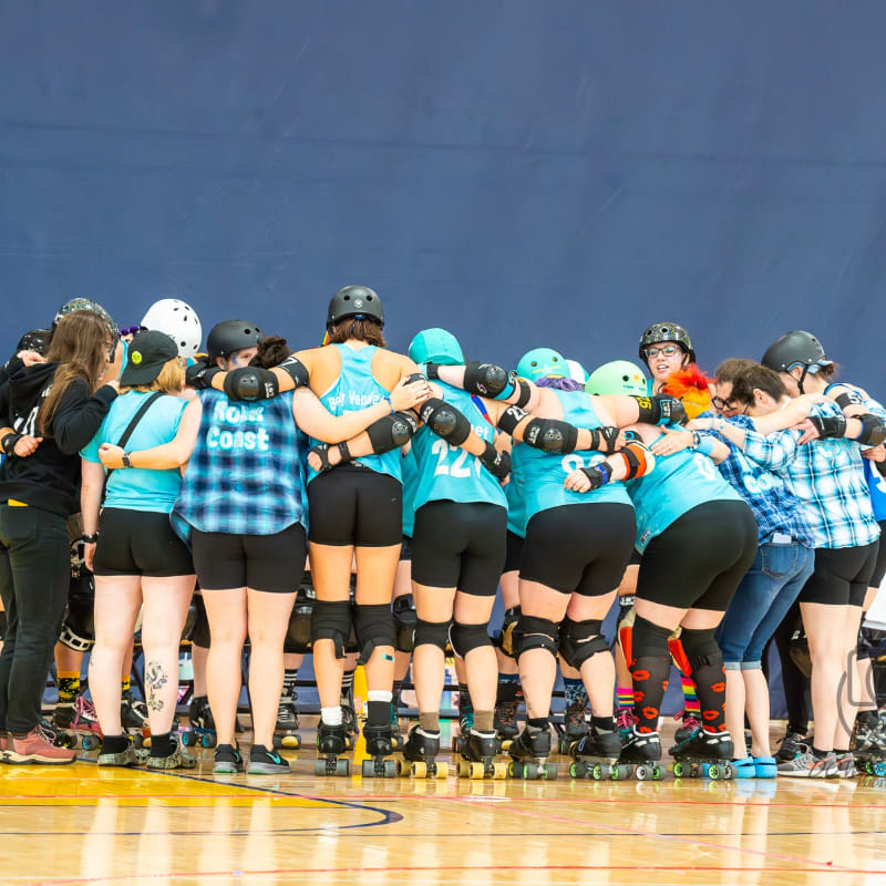 A group of roller derby players in teal jerseys and plaid shirts huddle together with their arms around each other in front of a blue wall.