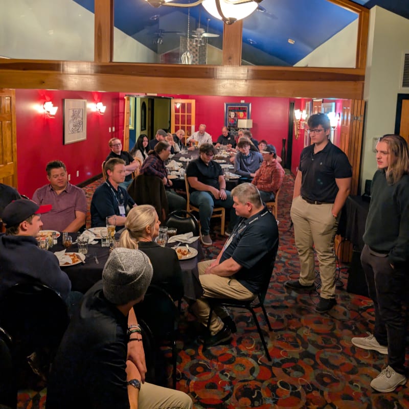 In the foreground, people are seated at round tables covered with black tablecloths, while in the background, a group of men stand near a doorway under a blue ceiling.