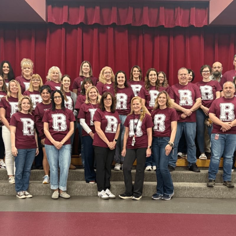 A group of adults in maroon t-shirts with a white "R" stand on risers in front of a maroon curtain.