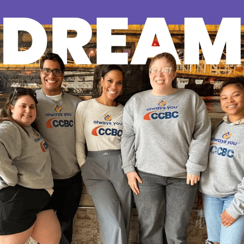 Five smiling individuals in matching CCBC sweatshirts stand in front of a brick wall with the word "DREAM" emblazoned above them.
