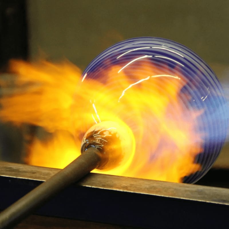 A molten glass orb on a blowpipe is being heated by flames against a blurred background of a workshop.