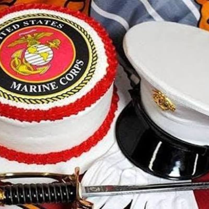 A U.S. Marine Corps cake and a white dress hat sit on a surface with a sword in the foreground, against a blurred background of fabric.