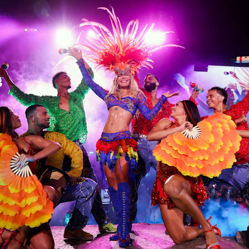 A vibrant carnival performer with a feathered headdress and colorful costume is center stage, surrounded by dancers with fans and a cheering crowd in the illuminated background.