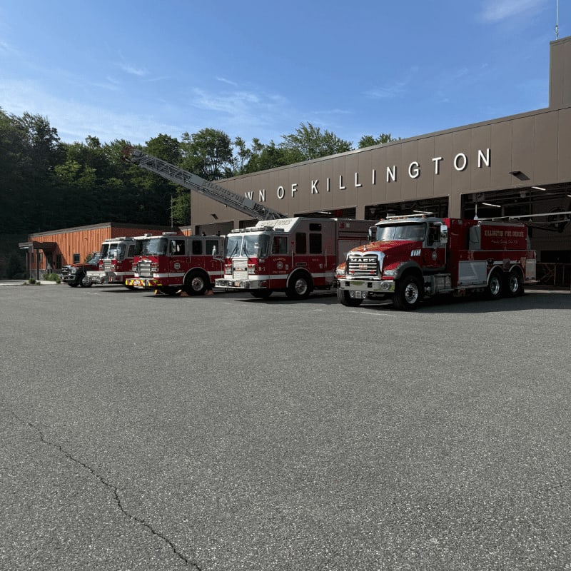Several red fire trucks are parked in front of the "Town of Killington" fire station, with a ladder extended from one of the trucks against a clear blue sky.