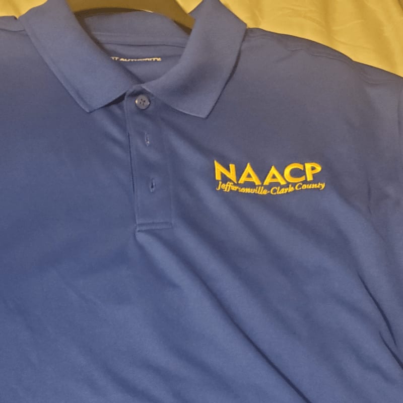 A blue polo shirt with the NAACP logo is laid out on a textured yellow surface.