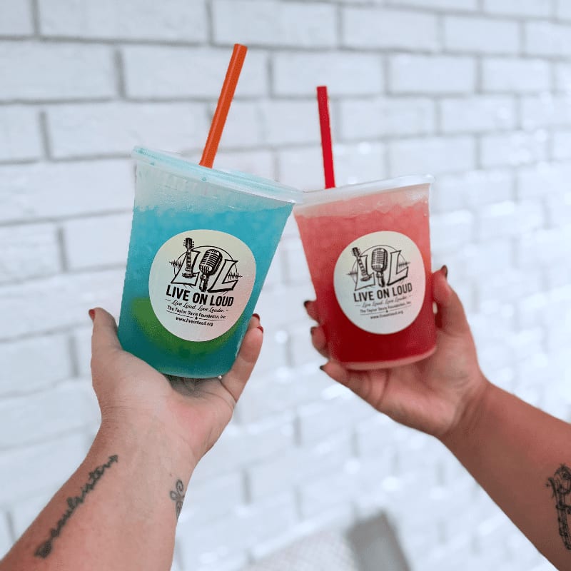 Two hands hold up colorful drinks with straws in front of a white brick wall.