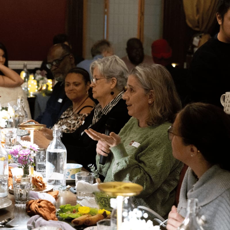 In the foreground, people are seated at a long table laden with food and drinks, while in the background, other guests are visible in a dimly lit room.