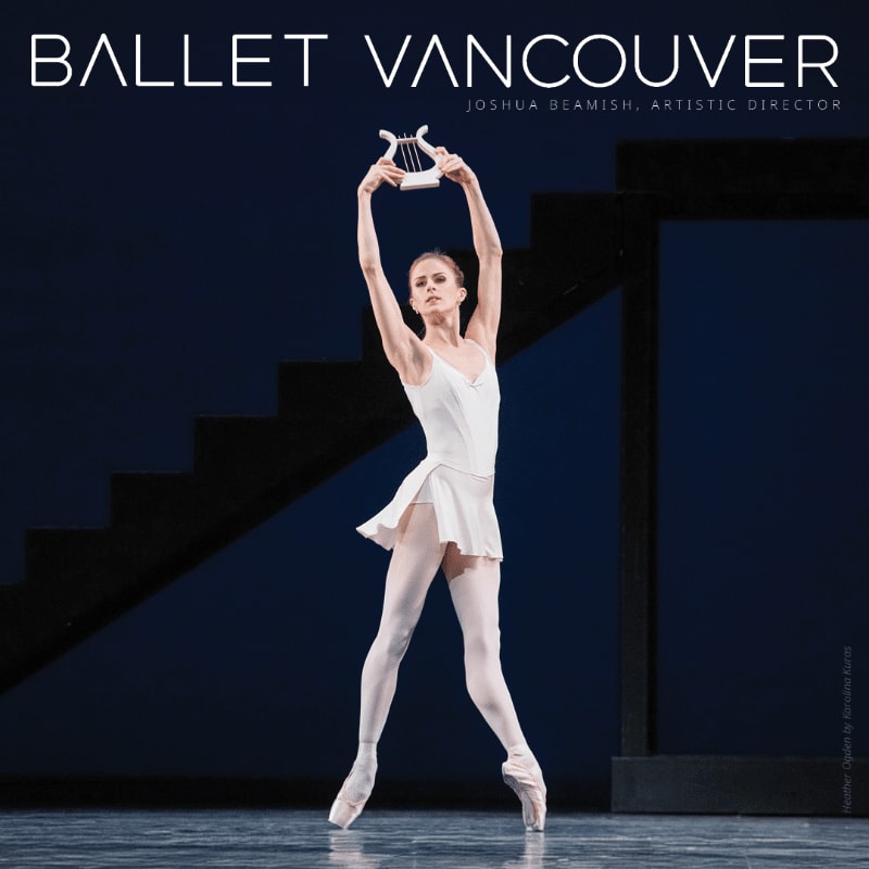 A ballerina in a white costume and pointe shoes holds a lyre above her head on a stage with a dark blue backdrop and a staircase.