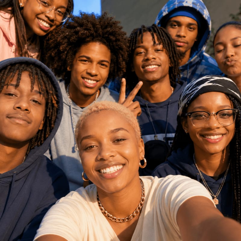 A group of smiling young people, some with their arms around each other, pose for a selfie with a blurred outdoor background.