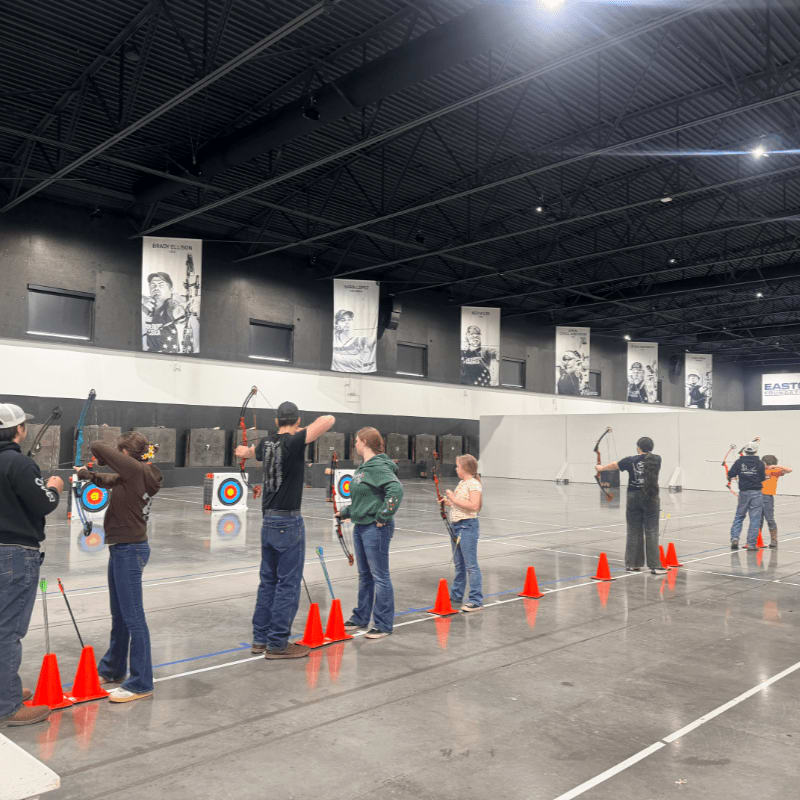 Several people practice archery indoors, with targets and banners visible in the background.