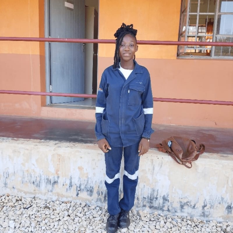 A young woman in a blue uniform stands on a gravel path in front of an orange building with a metal railing.
