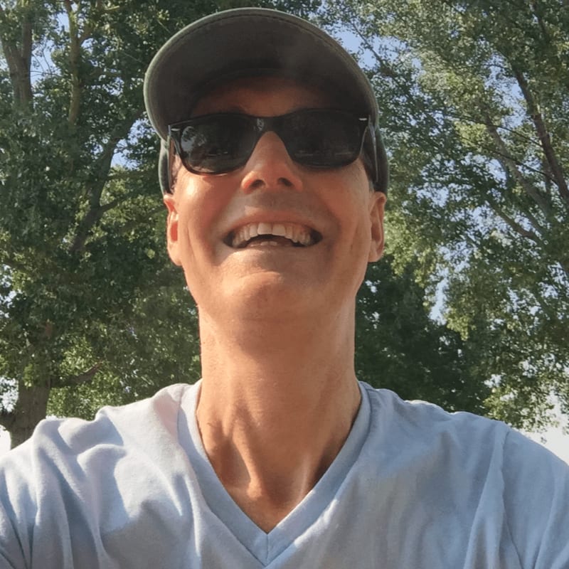 A man in a baseball cap and sunglasses smiles broadly in the foreground, with lush green trees and a bright sky in the background.