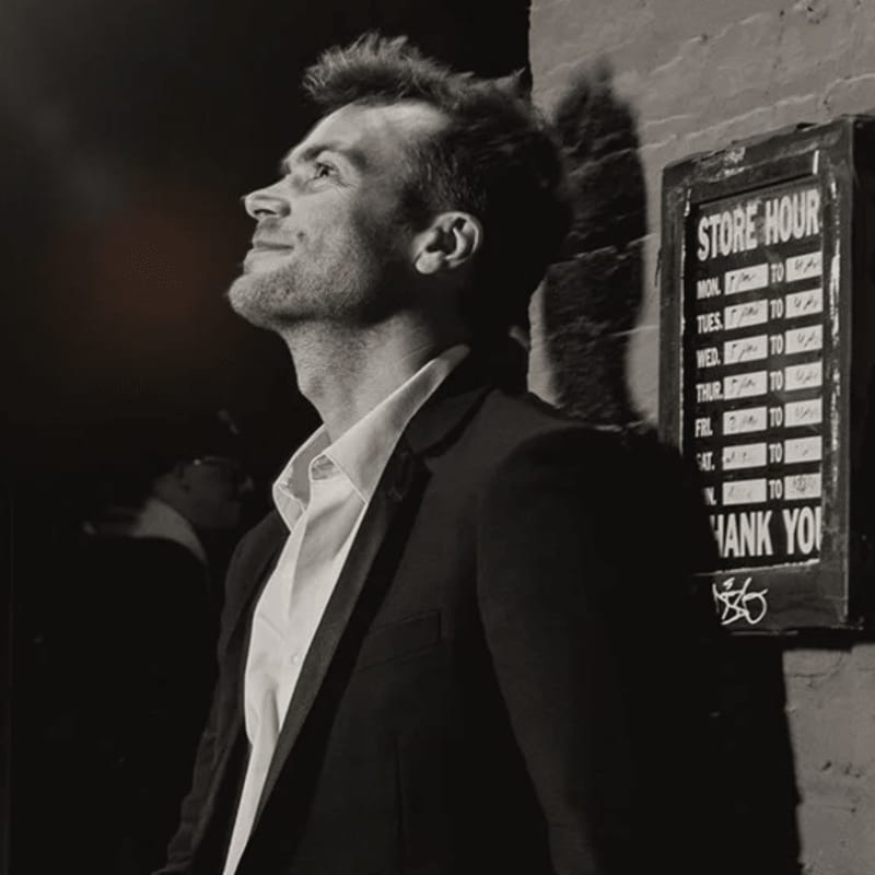A man in a suit looks up towards the sky in front of a brick wall with a "Store Hour" sign.