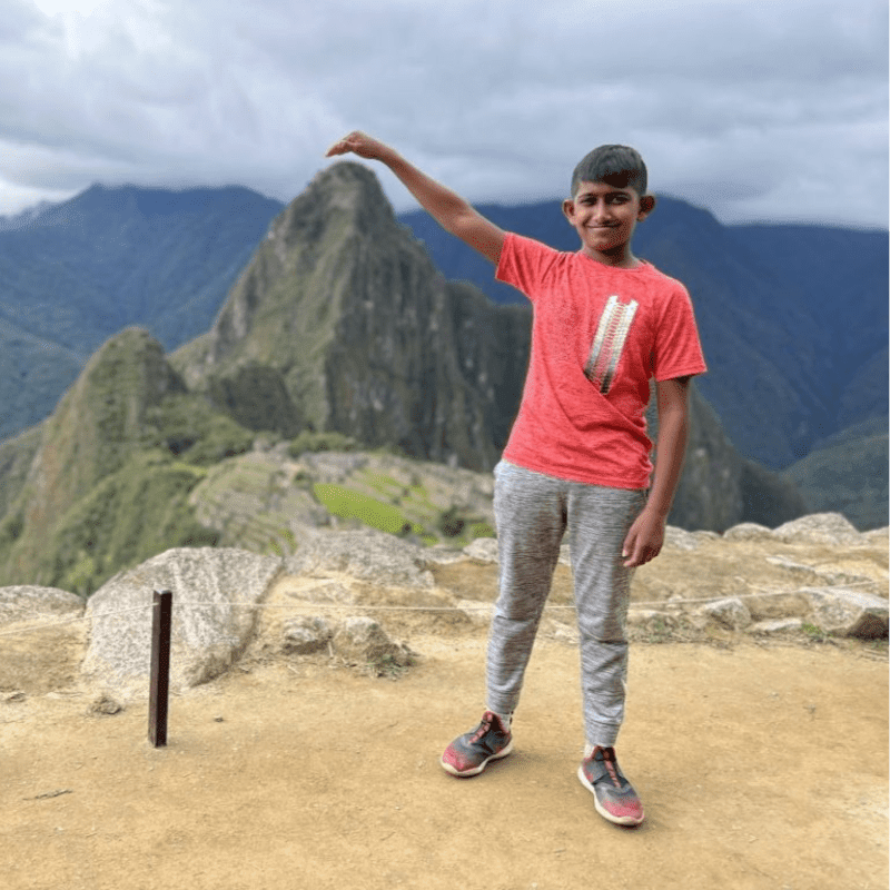 A young boy in a red shirt and gray pants stands on a rocky outcrop in the foreground, with the majestic Machu Picchu ruins and misty mountains in the background.