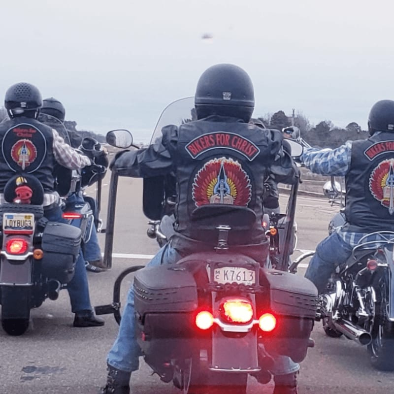 A group of motorcyclists wearing "Bikers for Christ" vests ride in formation on a road with a cloudy sky in the background.