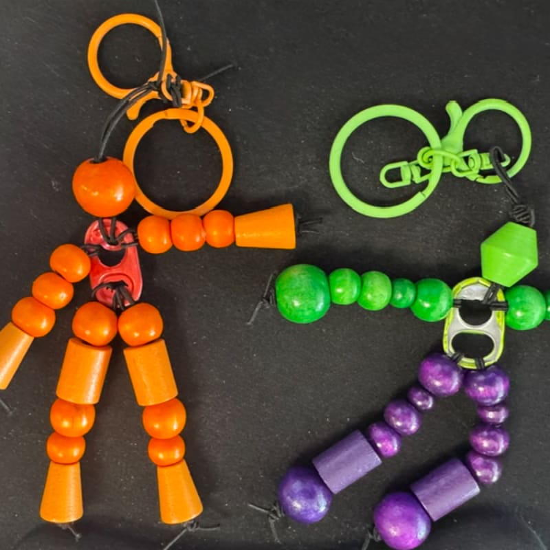 Two colorful, beaded keychain figures, one orange and red and the other green and purple, are laid out on a dark, textured surface with a bright green keychain loop in the background.