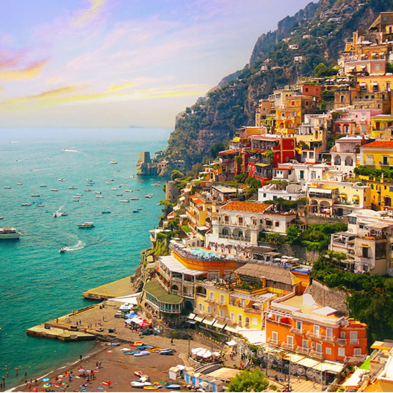 Colorful buildings cascade down a steep cliffside towards a bustling beach and a turquoise sea dotted with boats.