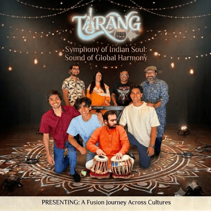 A group of musicians poses on a decorative floor with tabla drums in the foreground, while a banner with the event title "TARANG" hangs in the softly lit background.
